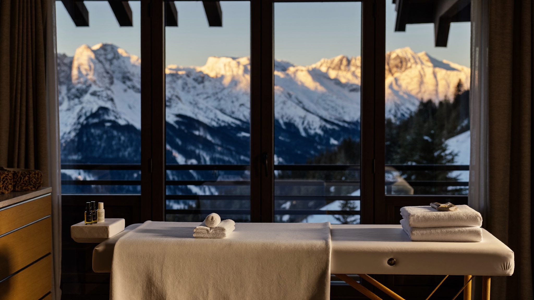 Massage table set before floor-to-ceiling chalet windows overlooking the snowy peaks of Les Arcs